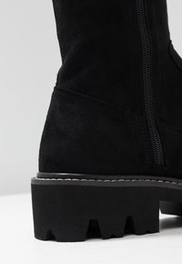 Botte noire en daim montant jusqu'au genou avec une semelle épaisse et texturée ainsi qu'une fermeture éclair latérale. Présente des coutures visibles le long de la semelle et une silhouette moderne.