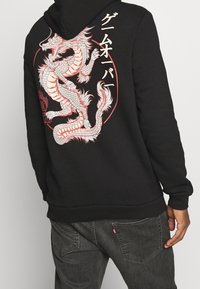 Czarna bluza z kapturem z dokładnym białym i czerwonym motywem smoka na plecach, z japońskim tekstem obok wzoru. Miękka tekstura materiału.