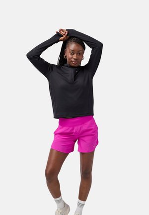 Schwarzer Pullover mit langen Ärmeln, Viertel-Reißverschluss und strukturiertem Stoff, kombiniert mit rosa Sportshorts. Das Model posiert mit erhobenen Armen.