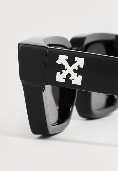 Gros plan sur des lunettes de soleil noires à monture épaisse avec un logo de flèches croisées blanches sur la branche.