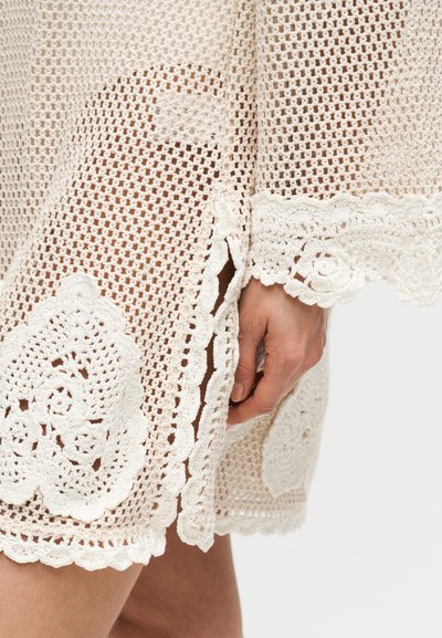 Χέρι που ακουμπά δίπλα σε μπεζ πλεκτή φούστα με κυματιστά άκρα και λουλουδάτα δαντελένια σχέδια.