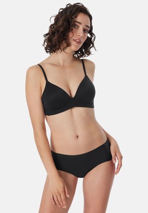 Femme aux cheveux bouclés posant en soutien-gorge noir sans coutures et sous-vêtements noirs assortis sur un fond clair uni.