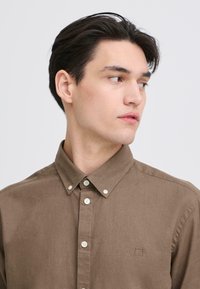 Chemise marron à boutons en tissu doux, avec un col boutonné, une surface texturée et un petit logo brodé sur la poitrine.