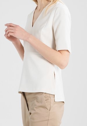 Eine Frau trägt eine weiße kurzärmlige V-Ausschnitt-Bluse, kombiniert mit beigen Hosen, und steht vor einem hellen, einfarbigen Hintergrund.