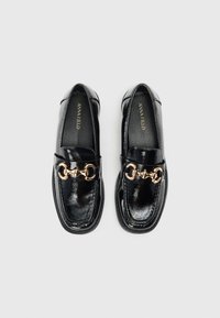 Mocasines negros de charol con un acabado brillante, suela plana y un acento de cadena dorada en la parte frontal. Etiqueta de tela visible en el interior.