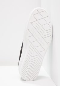Sneaker nera con suola in gomma bianca. La parte inferiore presenta un motivo a diamante e il logo in rilievo "Calvin Klein Jeans". Il lato ha dettagli in metallo.