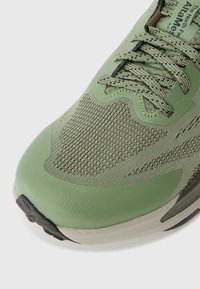 Chaussure de sport verte avec une tige en mesh texturé, un design épuré et une semelle en caoutchouc flexible. Comprend des accents et des coutures marron contrastants.
