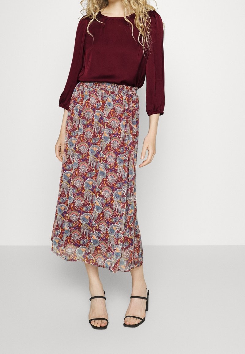 Femme portant un chemisier bordeaux à manches longues, une jupe midi à motif floral et des sandales noires à talons ouverts, debout sur un fond blanc.