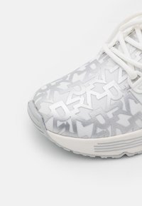 Weißer Sneaker mit überlappendem grauem stilisiertem Buchstabenmuster an der Spitze und weißen Schnürsenkeln auf einem hellgrauen Hintergrund.