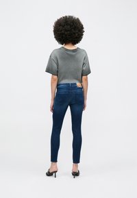 Kobieta o kręconych włosach ubrana w wyblakły szary T-shirt, ciemne obcisłe dżinsy i czarne wysokie mules, stojąca tyłem na białym tle.