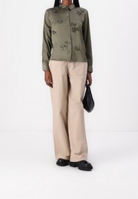 Camisa de satén verde oliva con adornos, diseño abotonado, mangas largas. Pantalones de pierna ancha beige, bolso negro, zapatos negros resistentes.