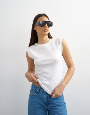 Frau mit schwarzen, übergroßen Sonnenbrillen, ärmellosem weißem Oberteil, blauen Jeans und mehreren Ringen, die vor einem schlichten hellen Hintergrund posiert.