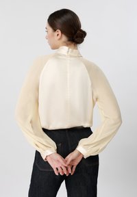 Kvinde med mørkt hår i en lav knold iført en cremefarvet satinbluse med gennemsigtige, lange ærmer, stående med hænderne foldet bag ryggen.