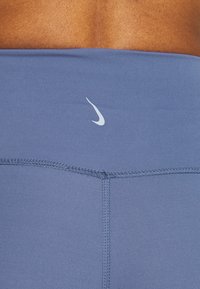 Detailní záběr dolní části zad osoby, která má na sobě modré sportovní legíny s malým bílým logem Nike ve tvaru swoosh umístěným na pásu.