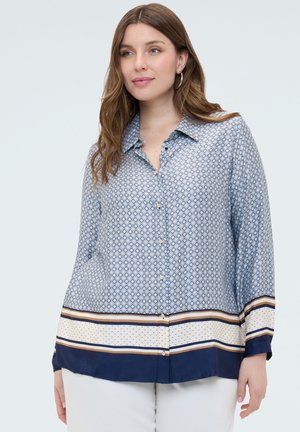 Frau trägt ein langärmliges Hemd mit blauen geometrischen und gestreiften Mustern kombiniert mit weißen Hosen.