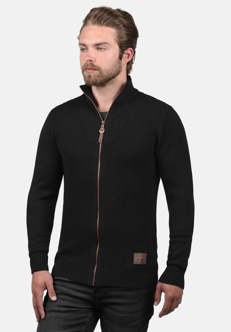 Homme aux cheveux bruns et à la barbe portant un pull noir à fermeture éclair avec un tricot texturé et un patch en cuir en bas à gauche.