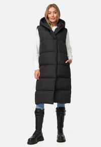 Svart vadderad väst med huva, med ärmlöst snitt, sidofickor och en strukturerad yta, bärs över en vit tröja och blå jeans.