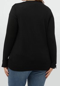 Žena v černém svetru s dlouhým rukávem a modrých джínách, zobrazená zezadu na jednoduchém světlém pozadí.