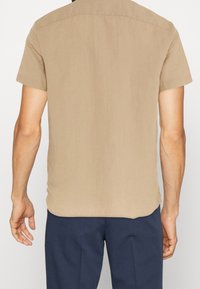 Camisa de manga corta color marrón claro, de corte recto, con una textura suave y detalles de costura sutiles en el hombro y la costura trasera.