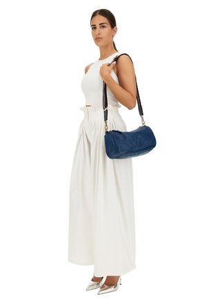 Borsa in pelle blu con una forma curvata, caratterizzata da una superficie testurizzata e una tracolla intrecciata. Modella che indossa un top corto bianco e pantaloni larghi.