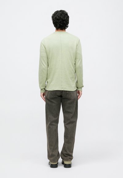 Šviesiai žalia ilgomis rankovėmis marškiniai su siūlėmis detale nugaroje, derinami su rudais tekstūruotais kelnėmis ir juodais bato, matoma iš nugaros.