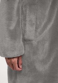 Grijze nepbontjas met een zachte textuur, voorzien van een zijzak en een gladde afwerking. De mouw heeft een subtiele manchetdetail.