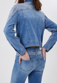 Nainen, joka käyttää vaaleansinistä farkkutakkia ja farkkuja, joissa on strassikoristelu olkapäissä, hihoissa ja vyötäröllä, kädet takataskuissa.