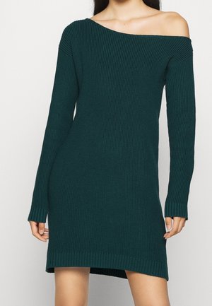 Femme portant une robe-pull côtelée vert foncé à épaules dénudées, manches longues et ourlet légèrement asymétrique.