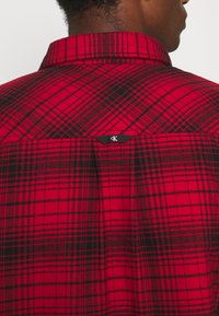Red and black plaid button-up shirt, featuring a collar, a tag with "CK," and a textured fabric with a structured fit.