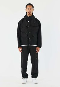 Giacca nera con cappuccio e multiple tasche frontali, abbinata a pantaloni cargo neri. L'outfit presenta una vestibilità comoda e sneakers bianche.