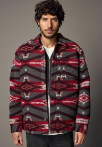 Veste en laine rouge et grise au motif géométrique, avec des boutons-pression et un col. Portée sur une chemise blanche.