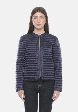 Donna che indossa una giacca trapuntata blu navy con cerniera e jeans scuri, in piedi contro uno sfondo grigio chiaro uniforme.