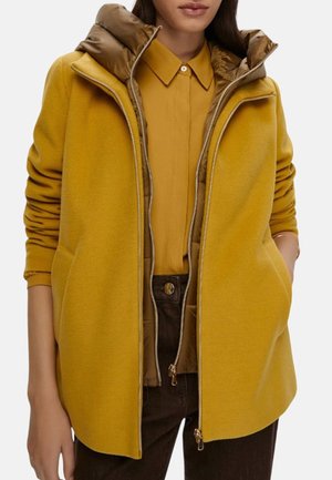 Cappotto giallo senape con cappuccio realizzato in tessuto testurizzato, tasche con doppia zip, foderato in contrasto e silhouette moderna. Abbinato a una camicia coordinata.