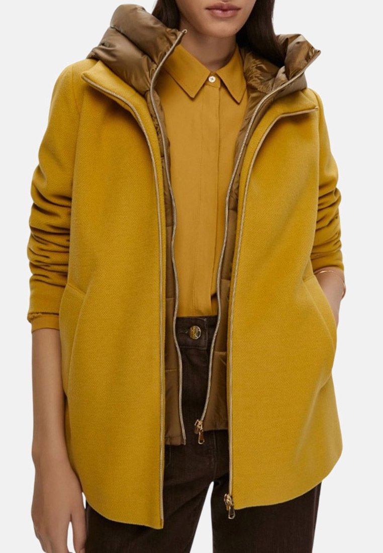 Cappotto giallo senape con cappuccio realizzato in tessuto testurizzato, tasche con doppia zip, foderato in contrasto e silhouette moderna. Abbinato a una camicia coordinata.