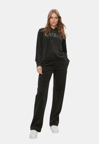 Tuta in velluto nero composta da una felpa con cappuccio con logo di strass e pantaloni abbinati dalla vestibilità comoda. Sneakers bianche completano il look.