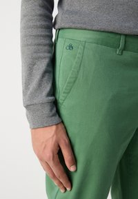 Grønne twillbukser med slim fit, sidelommer og et diskret broderet logo. Bæreren har en grå langærmet skjorte på.