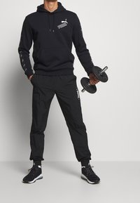 Felpa nera con logo sul petto e maniche stampate, abbinata a pantaloni joggers neri. Il modello tiene un manubrio e indossa scarpe da ginnastica nere.