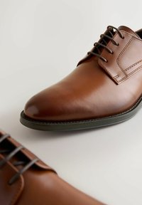 Brązowe skórzane buty do garnituru z wąskim noskiem, sznurowane, z subtelnym przeszyciem i kontrastującą ciemną podeszwą. Gładka tekstura, polerowane wykończenie.