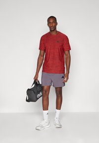Red, textured athletic t-shirt with black logo, paired with gray shorts. Holding a black gym bag with white logo, wearing white sports shoes.