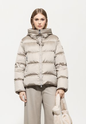 Frau in einer beigen Daunenjacke und Hose, die eine passende, weich strukturierte Handtasche vor einem schlichten hellen Hintergrund hält.