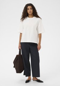 Hvid bomulds-T-shirt med korte ærmer, kombineret med mørkeblå vide bukser og sorte slip-on sko; holder en brun ruskindstaske.