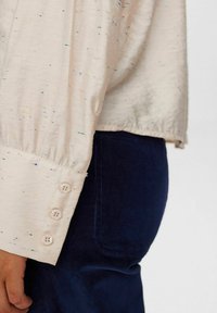 Gaiši bežs krekls ar krāsainiem plankumiem, ar pogājamiem piedurkņu galiem un brīvu piegriezumu, apvienots ar tumši zilu korduroja auduma biksēm. Gluda tekstūra un maiga auduma.