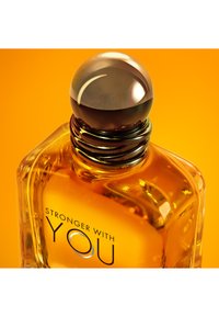 Bottiglia di profumo in vetro con liquido ambrato etichettata "Stronger With You" su sfondo arancione, con tappo metallico rotondo.