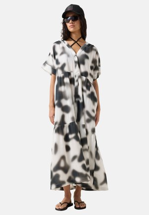 Woman wearing black and white abstract patterned maxi dress, black flip-flops, black cap, sunglasses, and a black strap necklace standing.