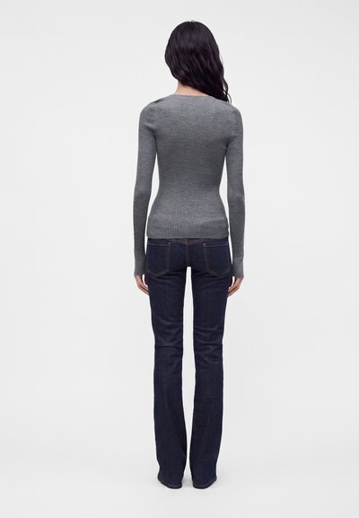 Femme aux longs cheveux foncés portant un pull gris côtelé ajusté et un jean bleu foncé, debout de dos sur un fond blanc uni.