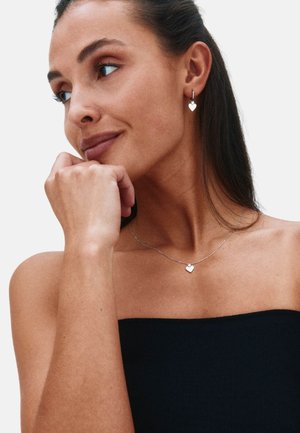 Silberne herzförmige Ohrringe und eine passende Halskette bei einer Person mit langen dunklen Haaren, die ein schwarzes trägerloses Oberteil trägt.