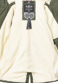 Fleece-lined outerwear featuring a cream interior, olive green sleeves and trim, with a black and white tag and decorative ribbon detail.