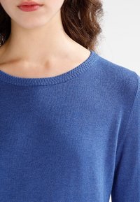 Femme portant un pull en maille bleu à col rond, montrant le cou, les épaules et le haut de la poitrine, avec des cheveux bruns bouclés partiellement visibles.