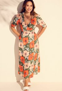 Blumenwickelkleid mit großen orangefarbenen und pinken Blumen auf grünem Hintergrund, kurzen Ärmeln und einem mittellangen Saum. Weiße Rutschsandalen.