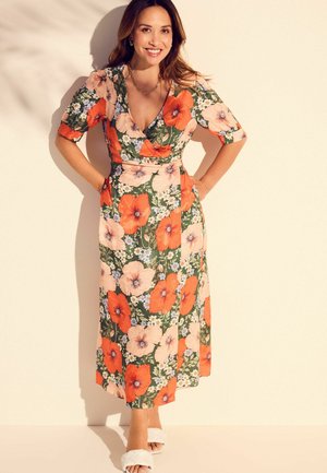 Blumenwickelkleid mit großen orangefarbenen und pinken Blumen auf grünem Hintergrund, kurzen Ärmeln und einem mittellangen Saum. Weiße Rutschsandalen.